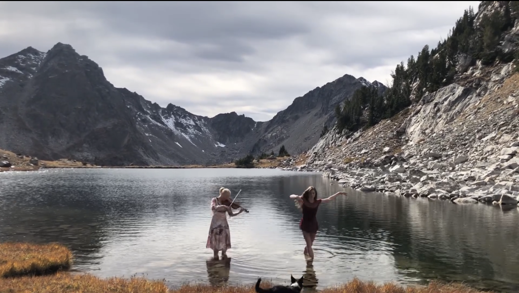 Two people stand in a mountain lake: one plays a violin, and the other dances. Snow-dusted peaks and rocky terrain surround them under a cloudy sky.